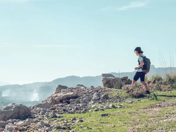 バックパック付きボーイは スペインのマヨルカの晴れた日にペレンサの町近くの山の尾根と青空に対する崖の端に向かって慎重に歩きます — ストック写真