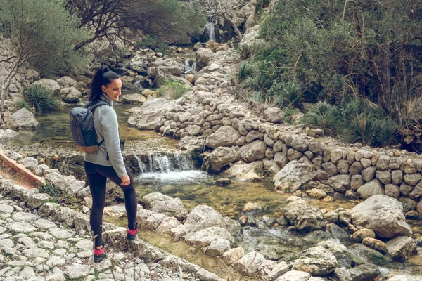 在Mallorca Soller Biniaraix 一个快乐的女人背着背包沿着狭窄的溪流瀑布 沿着清澈的小河走着 在岩石地带长满了绿色的灌木丛 — 图库照片