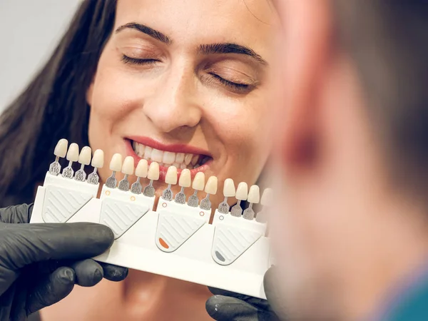 Dentist with palette of implants color choosing shade for veneers for young female sitting with closed eyes in dental office