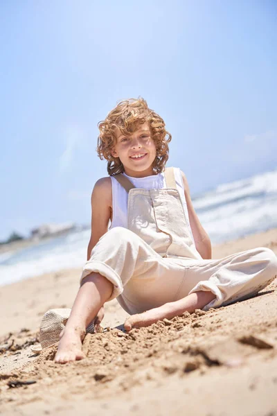 Full body of happy preteen kid with curly hair in jumpsuit sitting barefoot on seashore and looking at camera while enjoying summer holidays