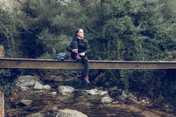 全身上下都是身穿马尾辫的积极的西班牙女徒步旅行者 他们躺在岩石河上的旧金属桥上 在马略卡岛的森林中跋涉时回头看去 — 图库照片