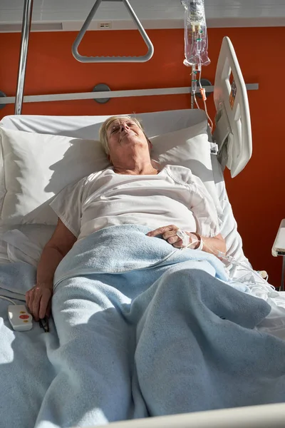From above of sick elderly female patient with short hair in white clothes lying on medical bed with connected venous catheter and sleeping during treatment in hospital