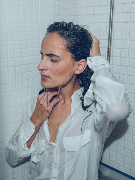 Thoughtful female in wet shirt standing with closed eyes under splashing water in shower cabin and touching head