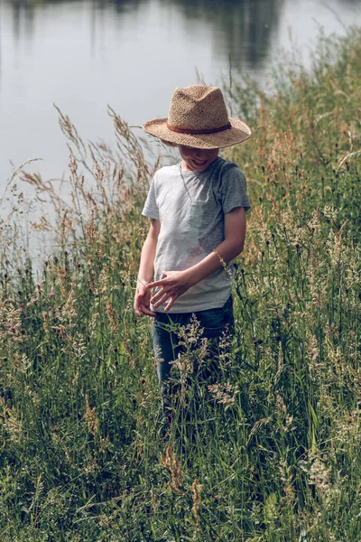 落ち着いたチャンネル近くの背の高い緑の草の間に立っているわら帽子で認識できない子供を笑い 下を見ながら夏の日を楽しむ — ストック写真
