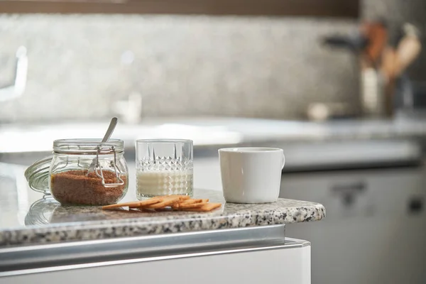 一杯热咖啡 牛奶和开胃饼干放在厨房里放红糖的玻璃瓶旁边的柜台上 — 图库照片