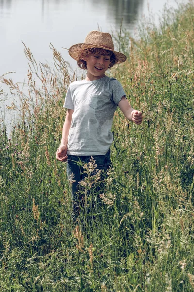チャンネル近くの背の高い緑の草の間に立っているわら帽子を身に着けているカーリー髪の幸せな子供 — ストック写真