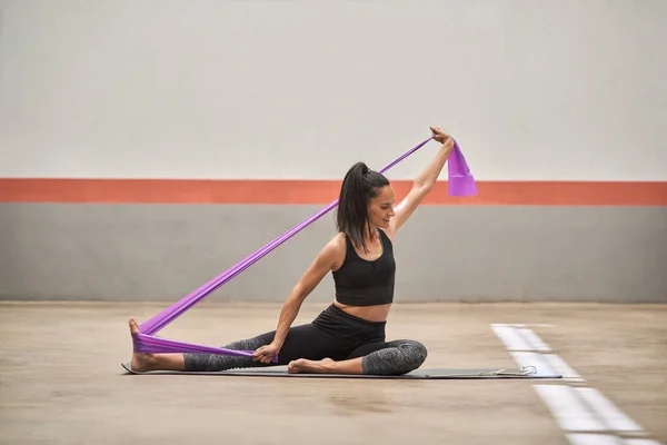 Full body of barefoot female athlete in sportswear sitting on mat and exercising with purple elastic band during training in fitness club while stretching legs and arms