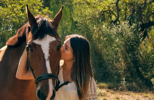 Gentle female equestrian kissing brown horse in muzzle while standing on meadow in summer forest