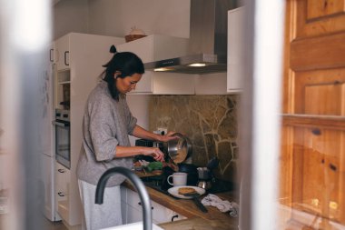 Mutfakta yemek pişirirken kaseden tavaya malzeme koyan ciddi bir kadının yan görüntüsü.