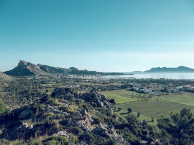 İspanya 'nın Mallorca kentindeki deniz kıyısında yeşil tarım arazileri ve dağların ortasındaki Pollensa kasabasının insansız hava aracı görüntüsü