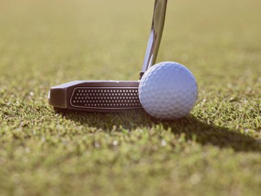 Metal siyah golf sopası, güneşli bir günde yeşil çimenlikte, beyaz, dengesiz bir golf topunun yanına yerleştirilmiş.