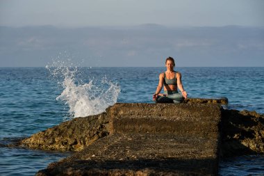 Padmasana pozu veren genç bayan sporcu. Elleri Gyan Mudra 'da kayanın üzerinde otururken ve deniz kenarında yoga yaparken.