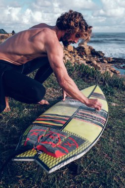 Tişörtsüz sörfçünün çimenli deniz kıyısına çömelmesi ve sörf tahtasında sörf yapmadan önce balmumu döşemesi.