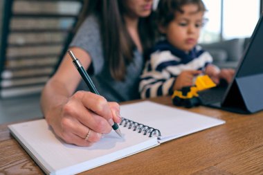 Tahta masada dizüstü bilgisayarla oturan ve küçük çocukla ilgilenirken not alan isimsiz bir kadın.