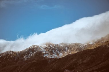 Pürüzlü dağ arazisi, doğada güneşli bir günde mavi gökyüzüne ve kalın beyaz bulutlara karşı beyaz toprağı kaplıyor.