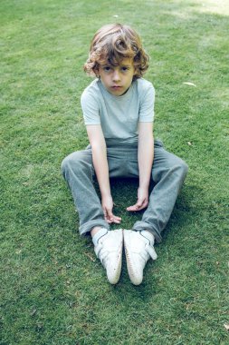 Kıvırcık sarı saçlı, yaz tarlasında yeşil çimlerde oturan ve güneşli bir günde kameraya bakan sevimli küçük bir çocuk.