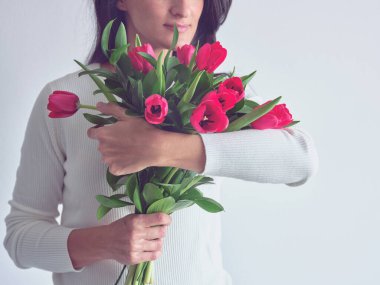 İsimsiz bir kadın beyaz arka planda dururken yüzünde hoş bir gülümsemeyle bir sürü laleye dokunup kokluyor.