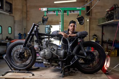 Ciddi sakallı, üniformalı bir tamirci parçalanmış motosiklete yaslanıyor ve eski püskü bir atölyede kameraya bakıyor.