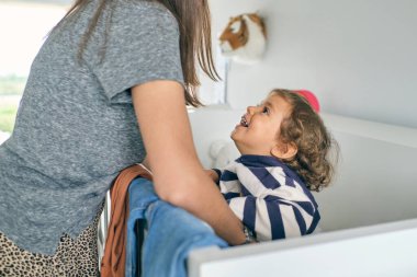 Beyaz bir karyolada duran, gülümseyen küçük çocukla oynayan ve evdeki odaya bakan kadının yan görüntüsü