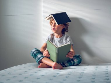 Bütün vücudu kitapla kaplı, pijamalı, açık bir kitapla yatakta oturan yorgun, sevimli bir çocuk.