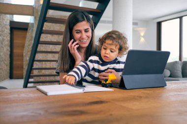 Oğluyla oturan, gülümseyen, tabletle ahşap masada oturan ve cep telefonuyla konuşan olumlu genç bir kadın.