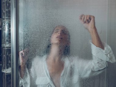 Duş kabininde polietilen perdenin arkasında dikilirken beyaz gömlekli, ıslak kadın gözlerini kapatıyor ve kollarını kaldırıyor.