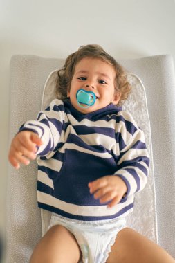 Mavi emzik takmış kaygısız küçük bir çocuk. Gülümsüyor ve kameraya bakıyor. Beyazların masasına uzanıyor ve evdeki ışık odasına uzanıyor.