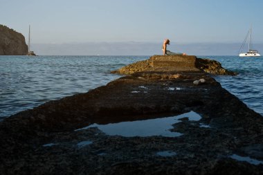 Aktif giyinmiş, Bhujangasana pozu almış, dalgalı mavi okyanusun yanındaki kayalık iskelede uzanan tanınmamış bayan sporcunun yan görüntüsü.