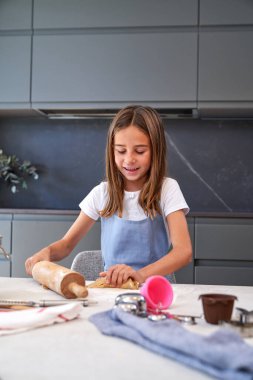 Evdeki hafif modern mutfakta pişirmeye hazırlanırken ahşap oklavayla kurabiye hamuru yoğuran önlüklü iyimser kız.