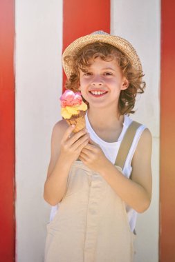 Hasır şapkalı mutlu çocuk gülümseyip kameraya bakarken kırmızı ve beyaz çizgili arka planda duruyor ve yaz günü waffle külahında dondurma yiyor.