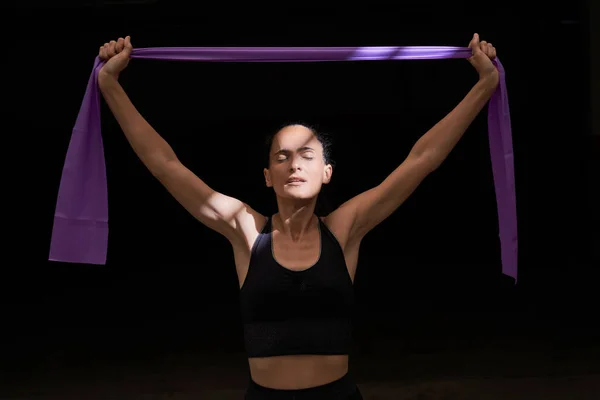 在黑暗的房间里训练时 穿着运动服的女运动员在脸上涂上太阳光 手臂展开 同时带着紫色的抵抗带运动 — 图库照片