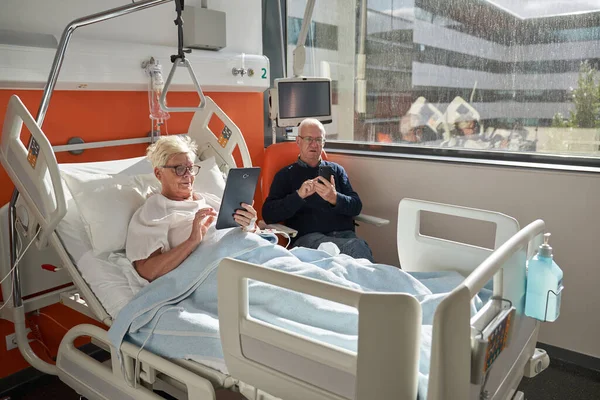 Full body of calm senior female patient with short hair in eyeglasses lying on medical bed and using tablet near husband messaging on smartphone in modern hospital