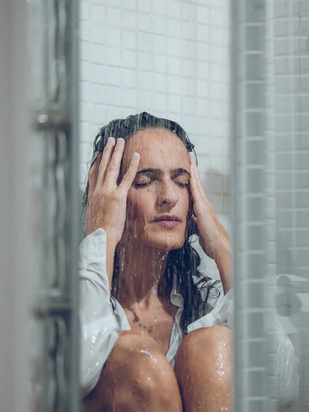 Depressed woman with dark hair in wet white shirt and with closed eyes sitting in shower under water stream and touching head gently
