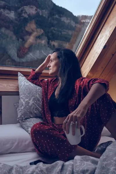 コテージの窓の近くに座って高地の森の景色を楽しんでいる居心地の良いパジャマの高級女性 — ストック写真