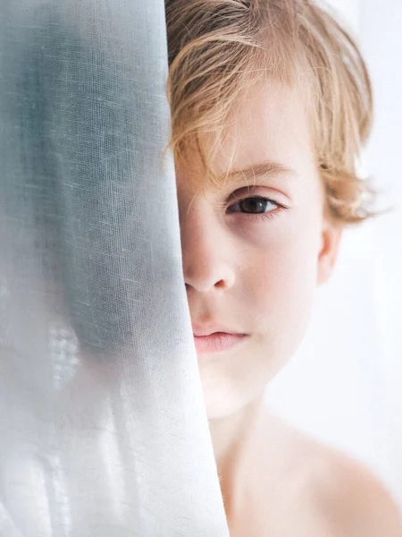ブロンドの髪と茶色の目がカーテンの後ろに隠れてカメラを見ている平和な少年 — ストック写真