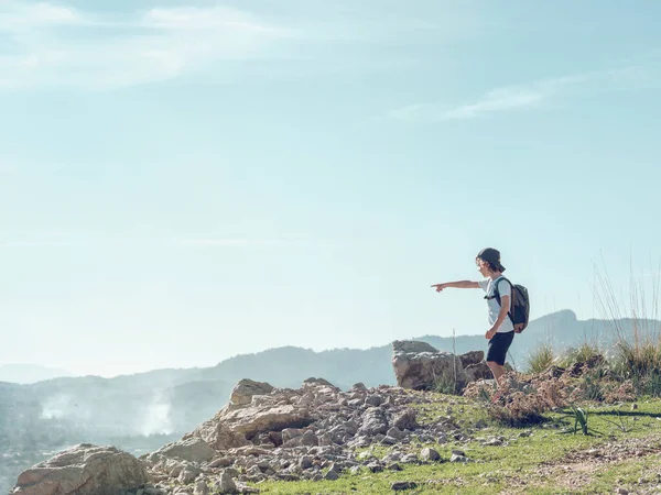 スペインのマヨルカの晴れた日に曇った青空の崖の上に立っている間 バックパックを指し 山の尾根を賞賛する子供の側面の景色 — ストック写真