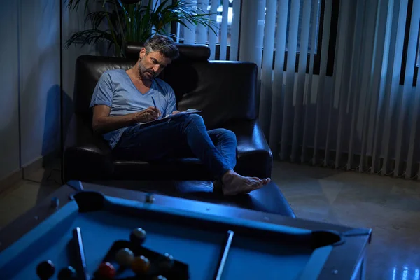Full length of businessman in casual clothes writing notes while resting in comfortable armchair in cozy office room at night