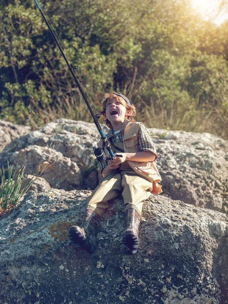 川の岩の岸に座っている間 釣り棒で魚を捕まえる開いた口を持つ幸せな男の子の全身 — ストック写真