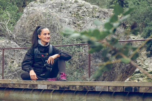 全身上下都是积极的西班牙裔年轻女性旅行者 她们身披着长长的黑色马尾辫 在索勒徒步旅行时 坐在年迈的桥上 面带微笑 望着远方 — 图库照片