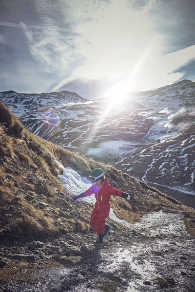 アウターウェアとヘルメットの子供は 雪の山の晴れた春の日に小さなクリークの近くを歩いている間 腕と岩でバランスをとります — ストック写真