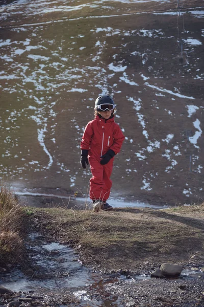 アウターウェアとヘルメットの子供は 高地で晴れた春の日を過ごしながら雪山の斜面に対して濡れた地面を歩いています — ストック写真