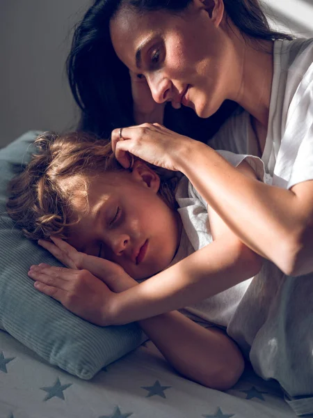 温柔的母亲把睡着的儿子的头发塞到耳朵后面 躺在舒适的床上 家里阳光灿烂 — 图库照片