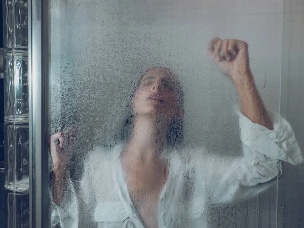シャワーキャビンのポリエチレンカーテンの後ろに立っている間 目を閉じて腕を上げる白いシャツのぬれた女性に苦しむ — ストック写真