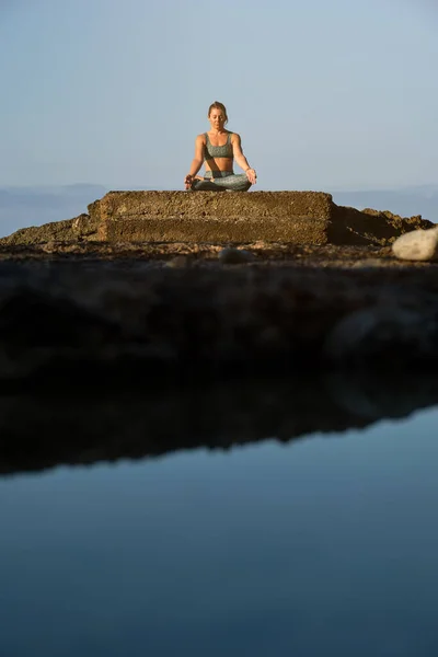 在蓝色海洋附近的巨石上打坐时 身穿运动服装的年轻女运动员手牵着手摆姿势 做着陀螺式的泥巴动作 — 图库照片