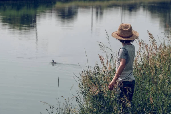 穿着草帽 站在水沟岸边绿草丛中 望着水面上的鸭子的难以辨认的孩子的侧视图 — 图库照片