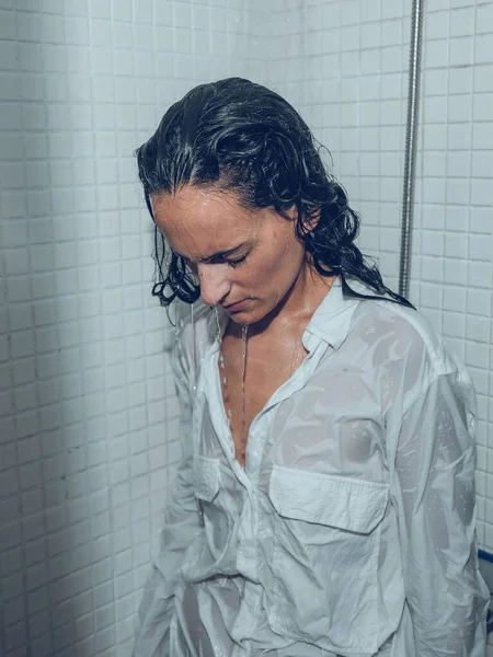 Tired woman in white shirt and with wet hair stranding with closed eyes and drooping head under spraying water in shower cabin and relaxing