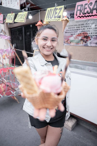 A young Hispanic woman is smiling holding a traditional ice cream in front of a street market stall. Concept of traditional Mexican dessert