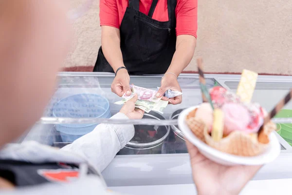 A young Hispanic woman is waiting for her change while holding a big traditional Mexican ice cream. Money selective focus. Concept of buying a sweet snack during summer