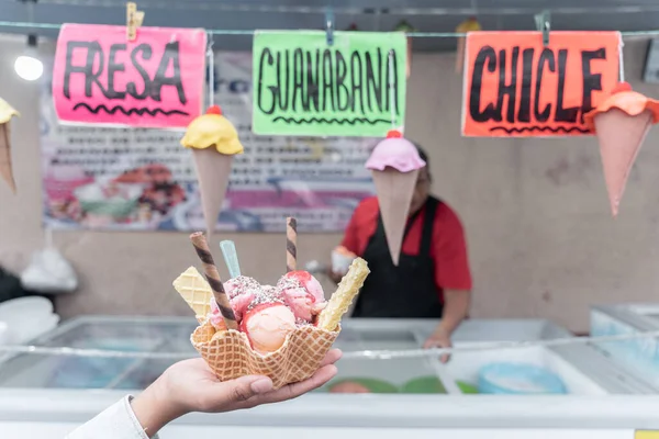 A hand is holding a traditional Mexican decorated ice cream in front of a street market stall with flavor signs. Concept of traditional Mexican dessert