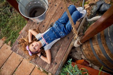 Sevimli bir çocuk vücudunun tamamından elleri başının arkasında, kırsal kesimdeki ahşap bankta dinlenirken yukarı bakıyor.